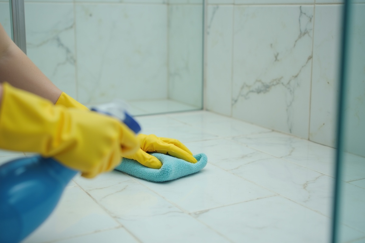 Person in rubber cleaning gloves wiping bathroom tile surface with sponge and cleaning spray, demonstrating proper bathroom cleaning technique near glass shower
